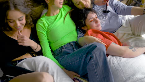 four women laying on bed while talk, drink and take pictures