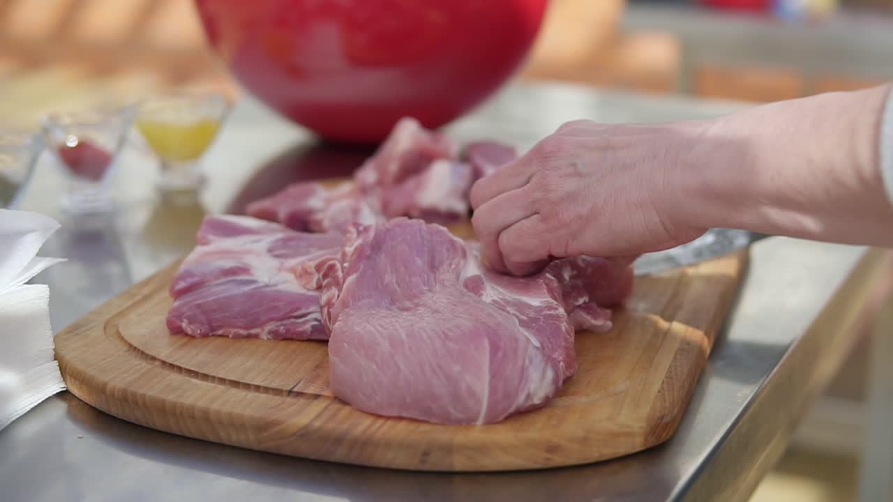 Person's hands cutting into a large piece of raw meat using a butcher's knife on a wooden surface outside. Barbeque preparation