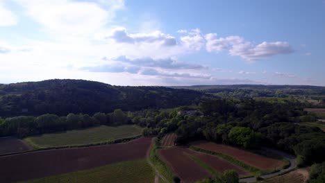 Luftaufnahme-Einer-Straße,-Umgeben-Von-Feldern-Und-Natur,-Vielen-Bäumen-Und-Grün,-Einem-Hügel-Auf-Der-Linken-Seite-Und-Dem-Horizont,-Blauem-Himmel-Mit-Wolken