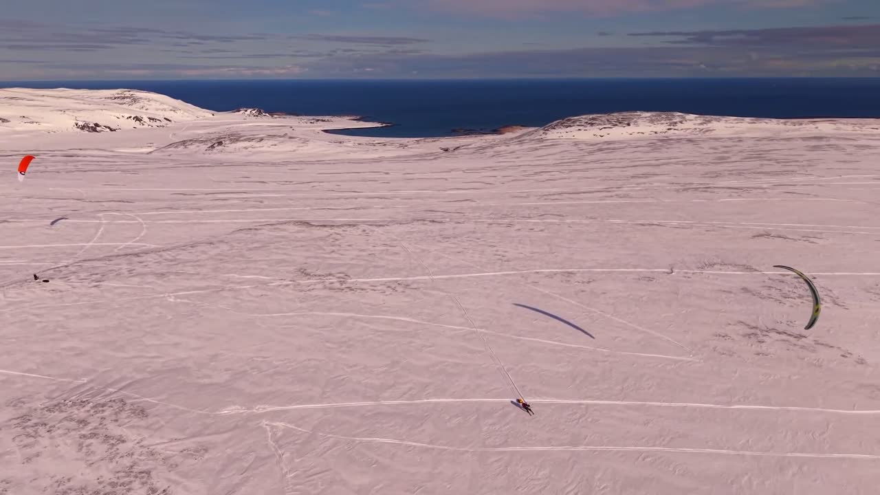 Aerial drone footage of people flying their kites and skiing on Norway Kite enduro or an endurance race during a beautiful sunny day in Norway while the Barents blue ocean sea water is visible in back