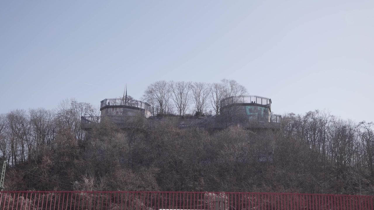 Una vista dal basso mostra le massicce strutture in cemento di una storica rovina di torre antiaerea che si erge su una collina boscosa, circondata da alberi invernali spogli sotto un cielo limpido e luminoso.