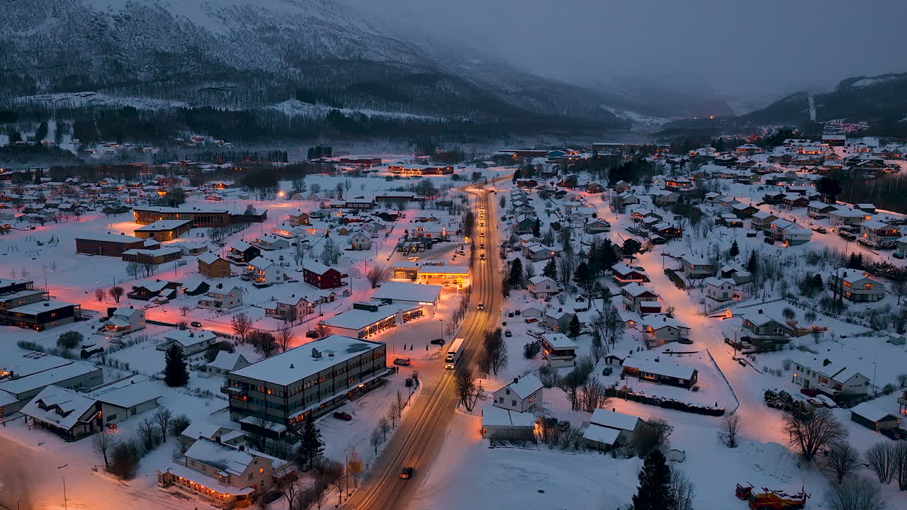 Ilmakuva pienestä norjalaisesta lumipeitteisestä kylästä Lofootien saaristossa sinisen tunnin aikana, hehkuvien katuvalojen ja liikenteen kanssa.