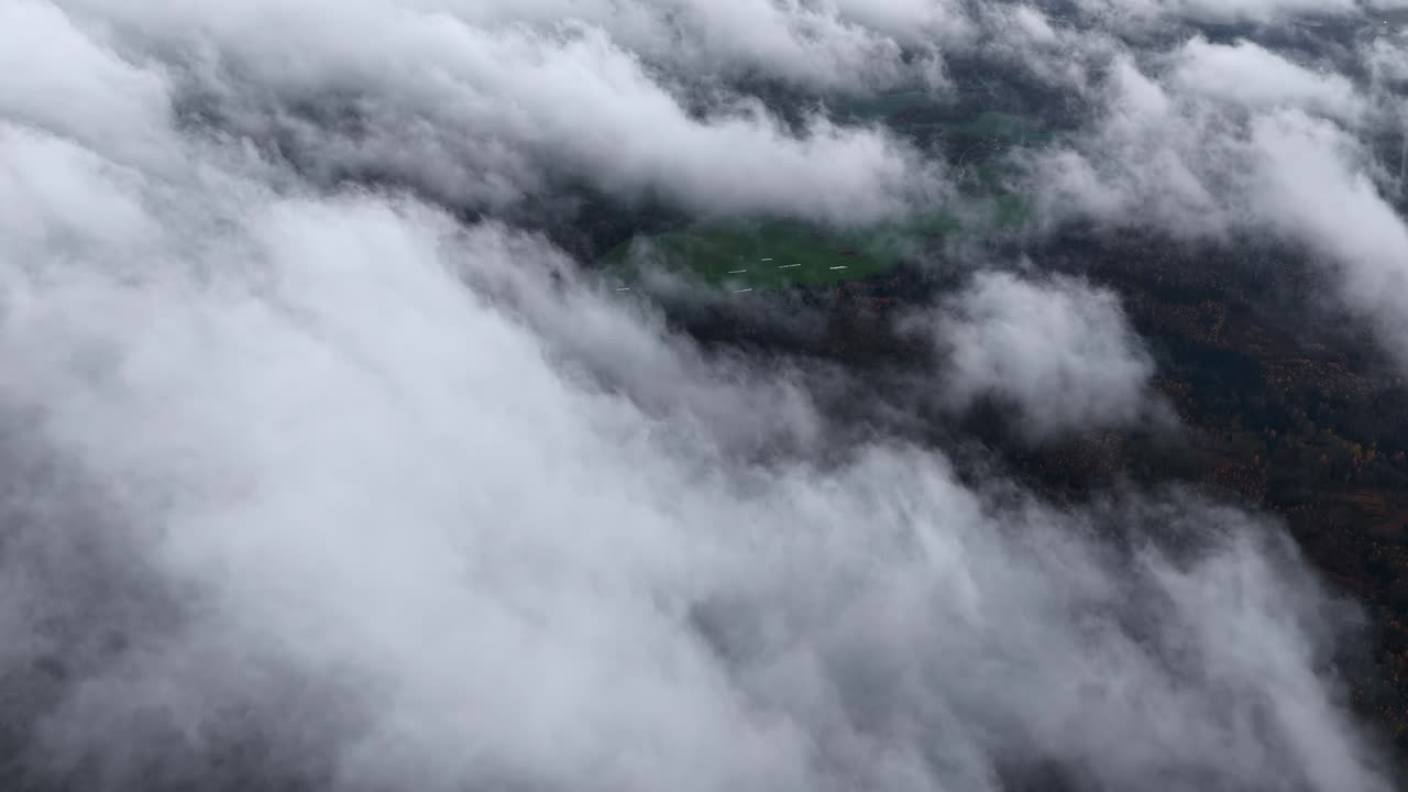 Aerial view above thick, low-hanging clouds partially revealing forest and farmland below