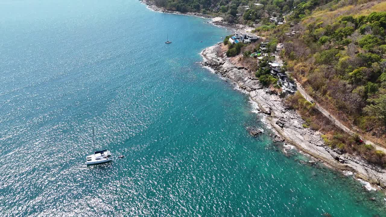 Drone footage captures a serene coastal scene in Phuket, Thailand, showcasing turquoise waters, rocky shores, and a sailing boat