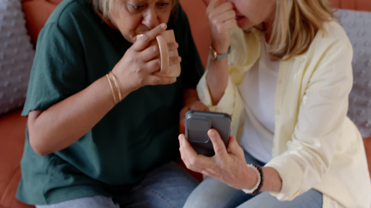 Holding smartphone, two multiracial senior female friends looking at screen and discussing, at home