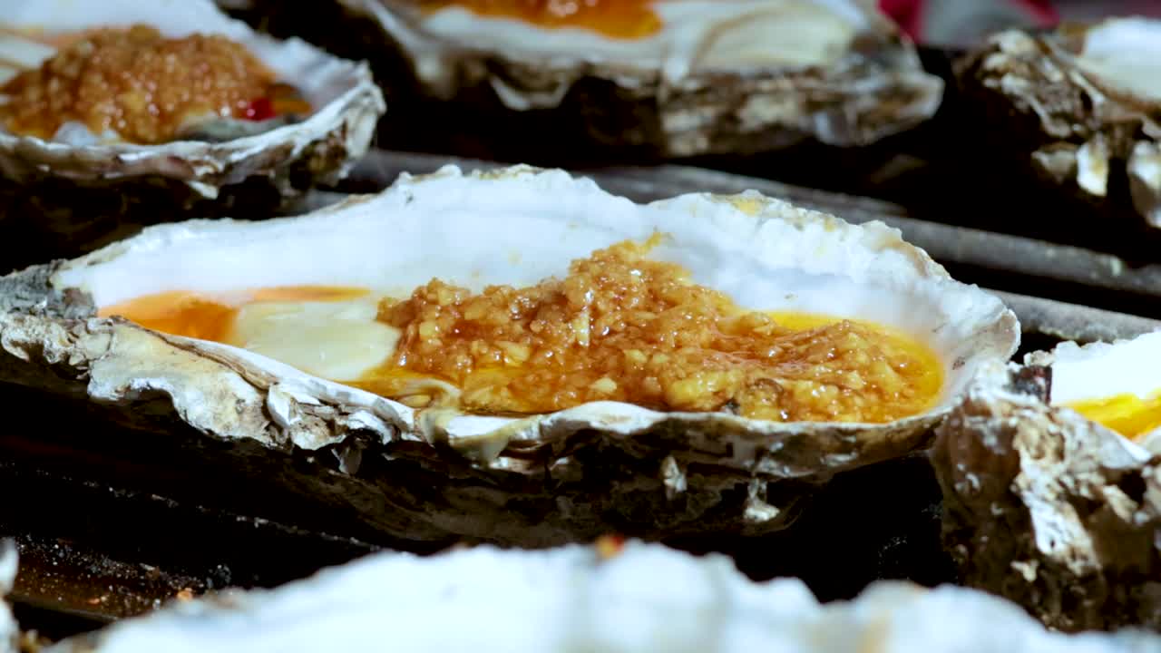 A detailed view of oysters being grilled, topped with garlic and sauce, highlighting their texture and preparation.