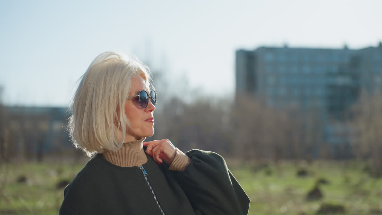 Confident blonde woman with bob hairstyle wearing sunglasses and dark coat standing outdoors in bright sunlight, holding collar of jacket, urban background with blurred buildings in distance