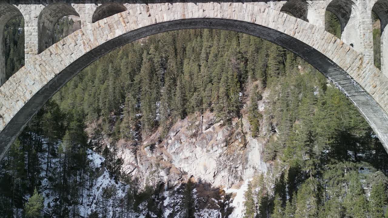 Drone passing by old stone arch bridge crossing deep gorge in Douro Valley, Portugal, showing pine trees, rocky cliffs, clear sky, and scenic landscape with forested terrain in background