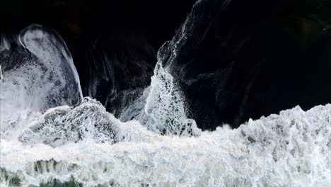 Aerial-birds-eye-view-of-white-ocean-tide-crashing-on-contrasting-black-sand-beach-in-Bali,-Indonesia