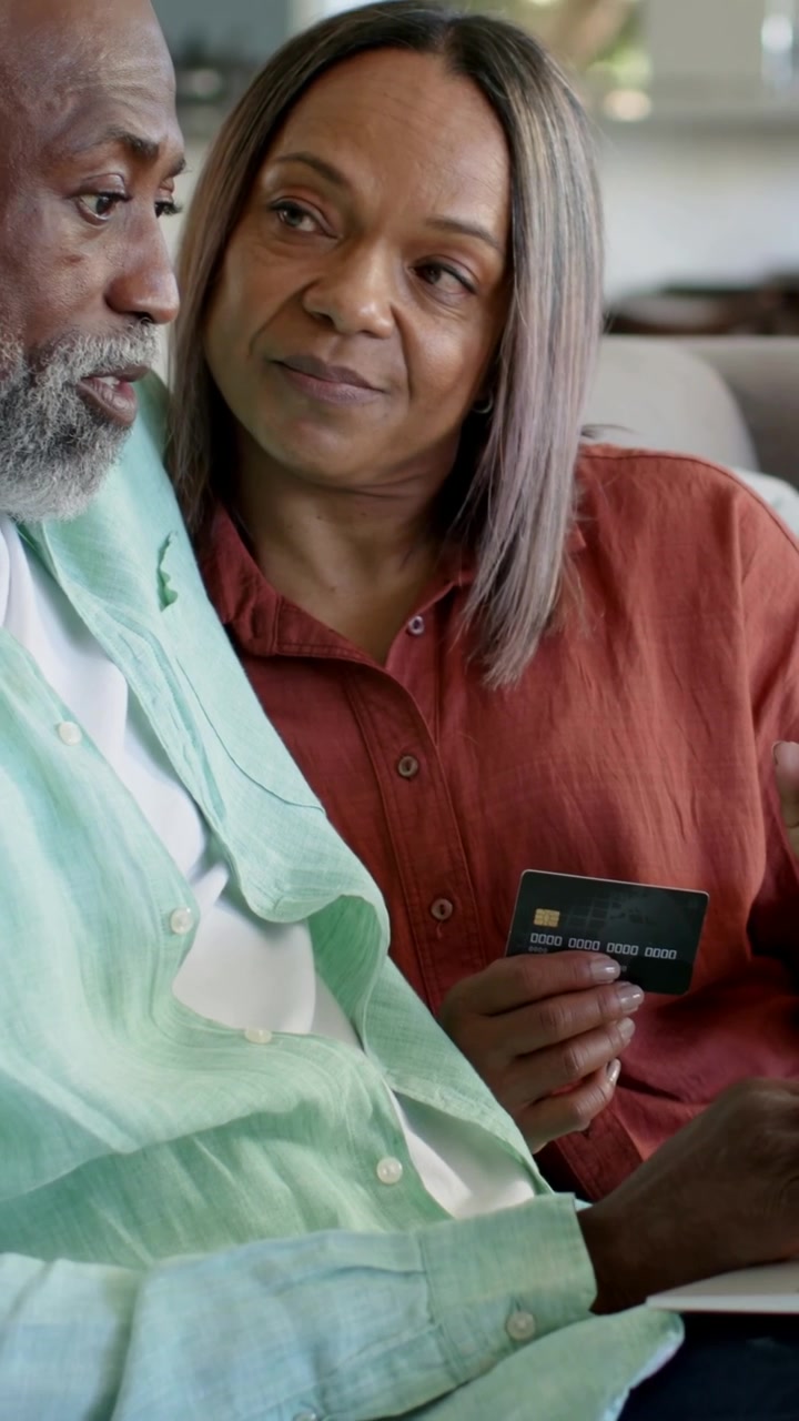 Vertical video: Holding card, rust shirt woman leaning to man tapping phone on couch, checking bill