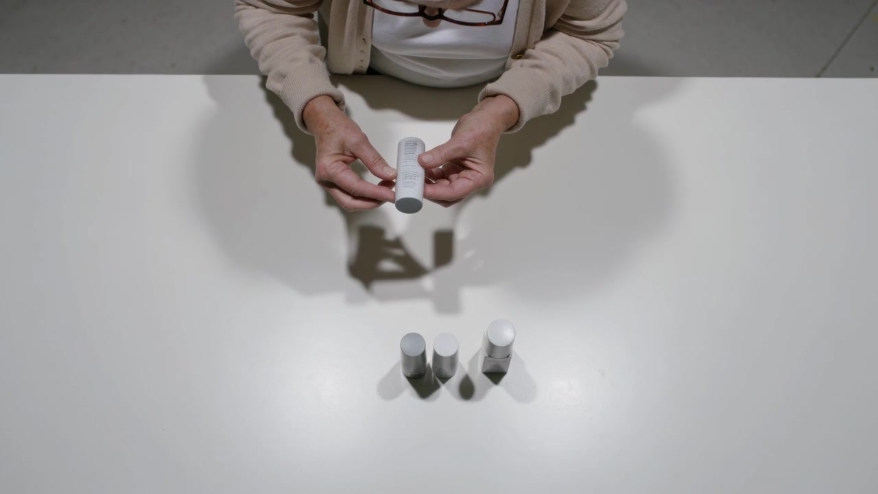 Grasping left bottle, senior woman in beige cardigan lifting and inspecting four bottles at table