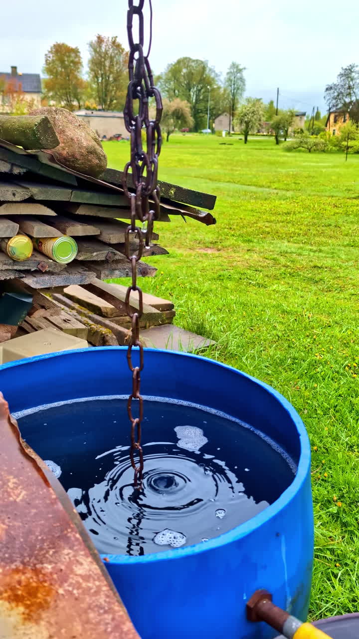 Rainwater drips down metal chain into blue barrel, drinkable water collection, vertical