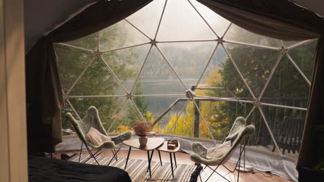 dining place on glamping glass terrace. modern hotel room for rent with panoramic windows to enjoy lake view on foggy autumn morning. relaxation space