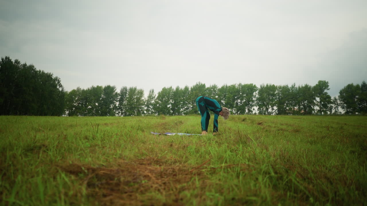 초록색과 검은색 의상을 입은 여성이 넓은 잔디에서 야외에서 구름이 가득한 하늘 아래에서 낮은 지 포즈를 연습하고, 나무가 배경에 줄지어 있습니다.