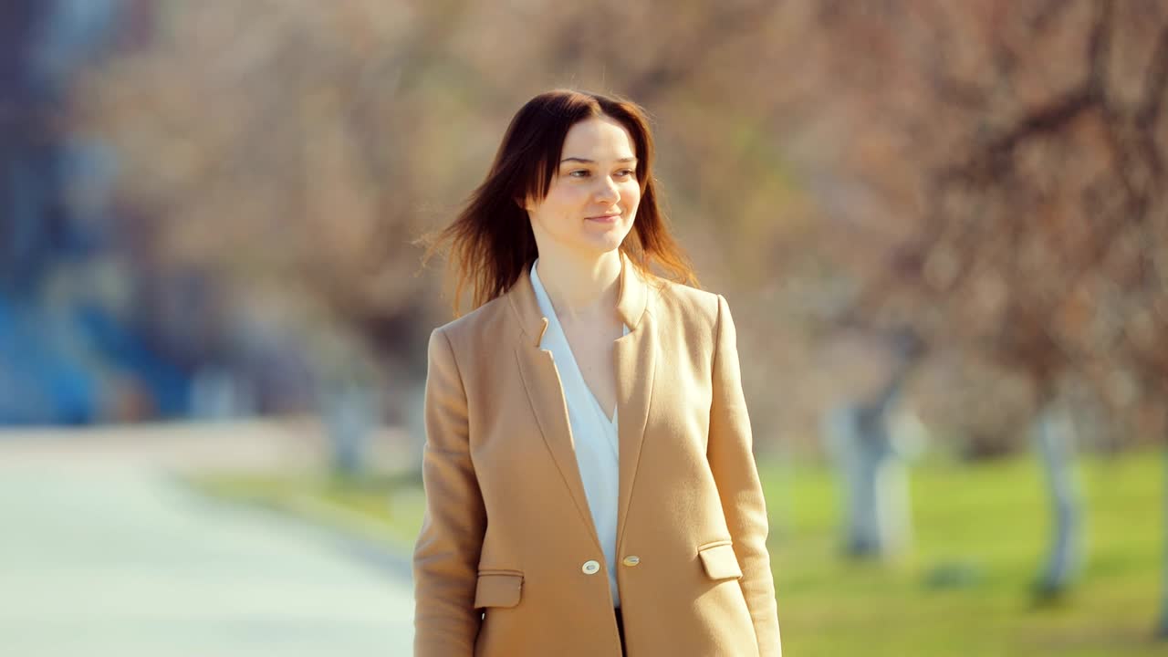 우아한 젊은 여자가 초봄에 공원을 걸어다니며 행복한 귀여운 여자가 광장에 가서 미소 짓는다.