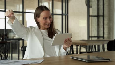 young woman happily enjoys winning using a tablet while sitting at a desk in the office