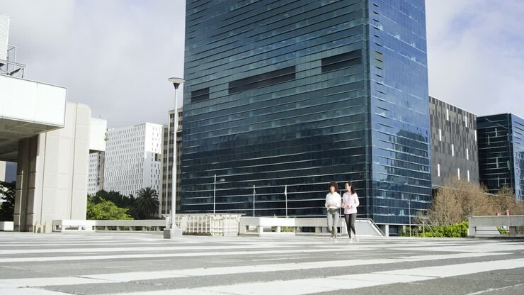 Duas amigas entrando em uma praça listrada, caminhando para correr sob uma torre de vidro azul, se exercitando