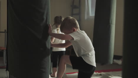 child attacks punching bag with classmates in gym. toddler boy beats hanging equipment by legs and hands in classroom slow motion. kickboxing team