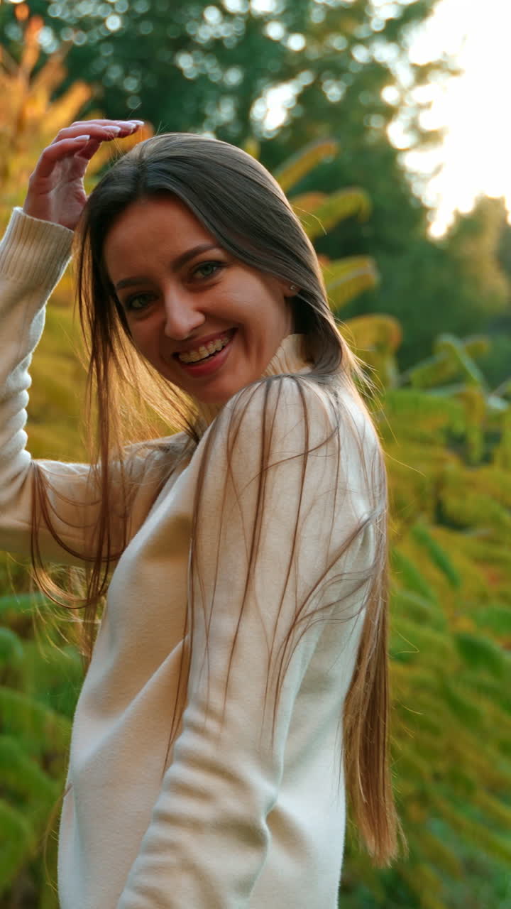 Happy smiling dark-haired woman waving her long hair in front of camera. Lady in white sweater posing at the backdrop of autumn park.
