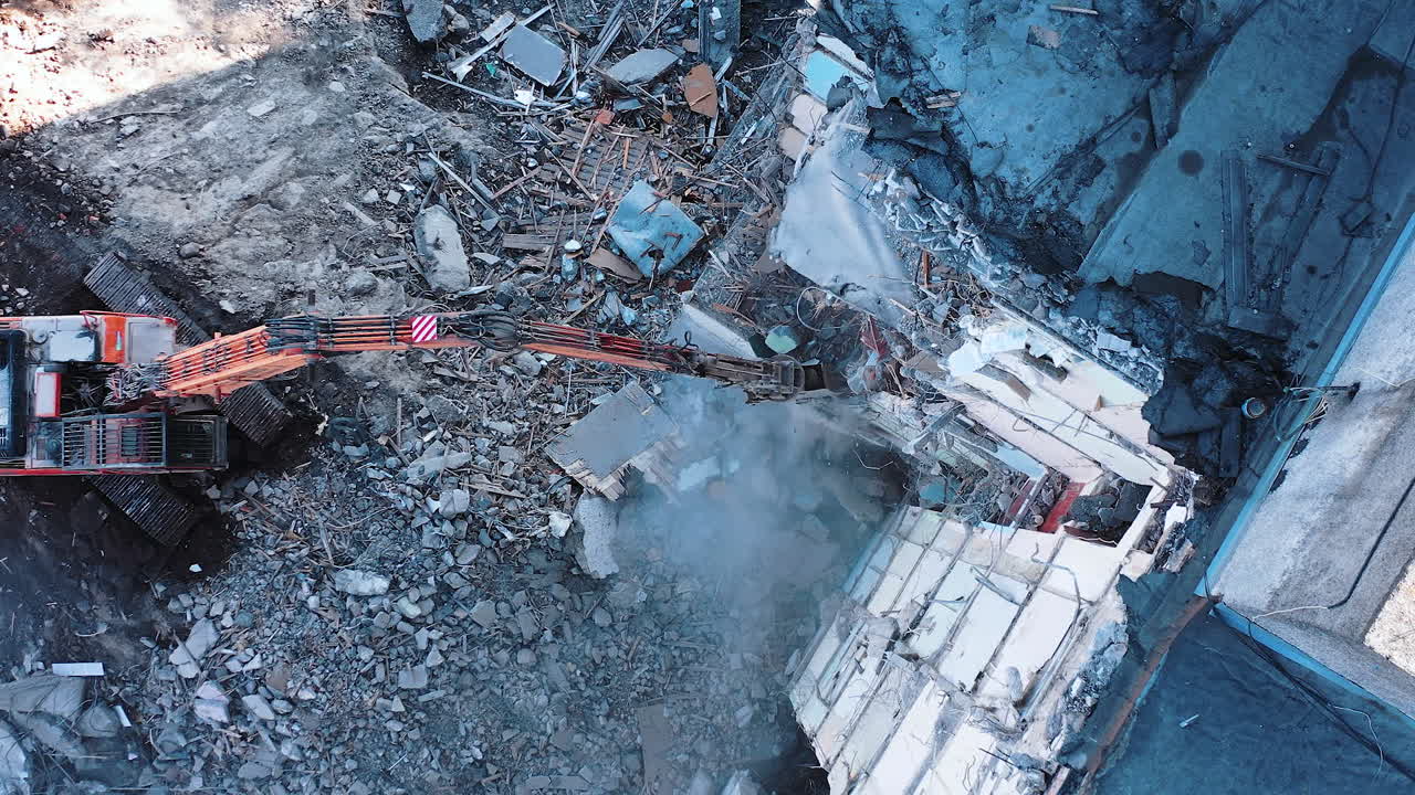Long claw of an excavator destroys the concrete parts of the building. Demolition of premises after an earthquake. Up view of excavators working with ruins of demolished buildings.