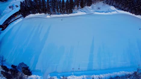 aerial rise over massive outdoor skating rink with2 people out during a twilight sunset lockdown covid19 with shadow reflections of tall pointy pine trees by public parks cabana onto the side left2-2