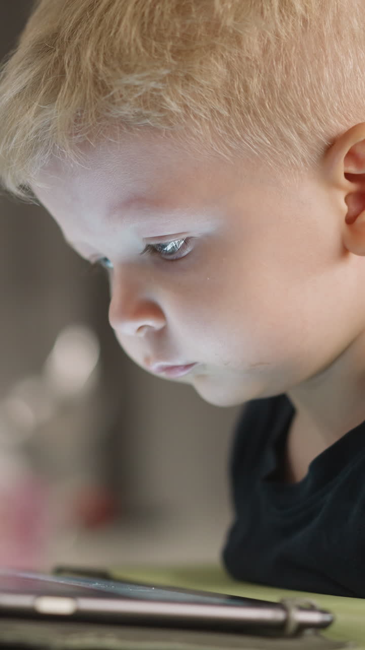 Blond toddler boy smiles while watches funny videos via tablet at home closeup. Cheerful little kid enjoys gadget. Internet technology of amusement