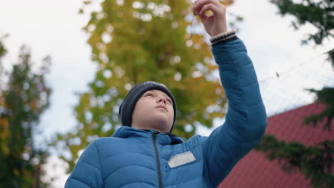 young boy in blue jacket and beanie holds dry leaf high, inspecting it carefully against backdrop of vibrant autumn foliage and rustic fence, exploring nature in outdoor setting