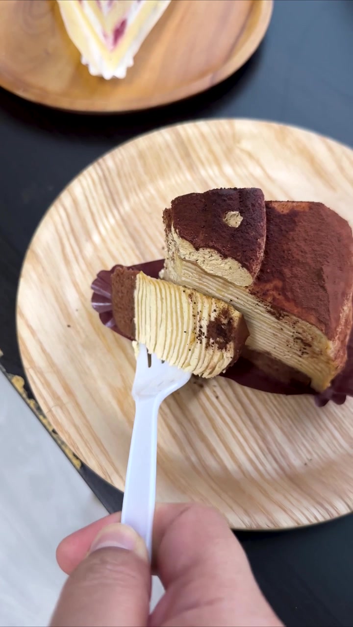 Doğal ışıkta, ahşap tabakta duran tiramisu dilimini plastik çatalla kesip kaldıran el.