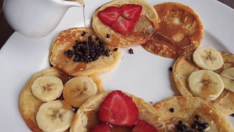 Vertiendo-Jarabe-De-Arce-En-Un-Movimiento-Circular-Sobre-Un-Elegante-Arreglo-De-Panqueques-Con-Rodajas-De-Fresas,-Plátano-Y-Chocolate