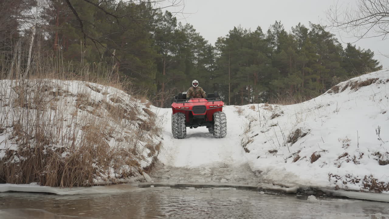 ATV rosso si lancia da un argine innevato in un torrente, il pilota è brevemente in aria prima di un drammatico schizzo, bordo d'acqua ghiacciato e ripido pendio di pini, un'ampia vista cattura la discesa e l'impatto, acrobazia fuoristrada ad alta adrenalina.