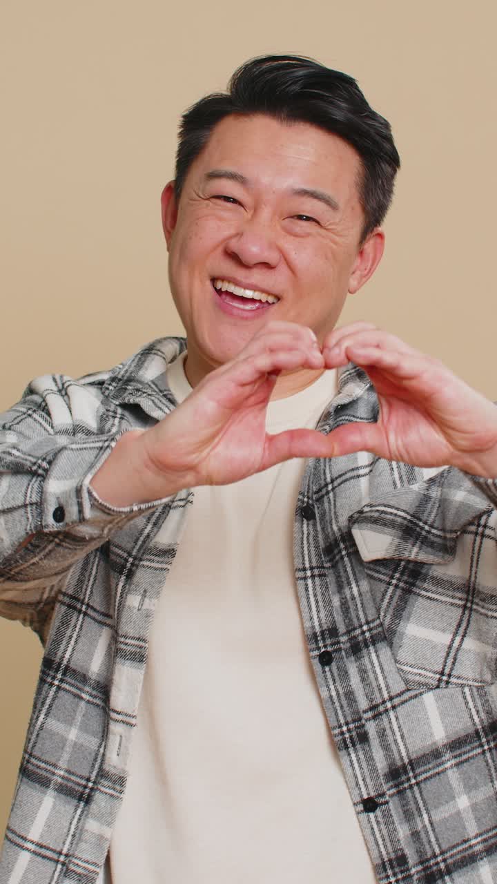 幸せそうなアジア人男性がハートのジェスチャーで愛情を示し、良い感情と共感を表現しています。