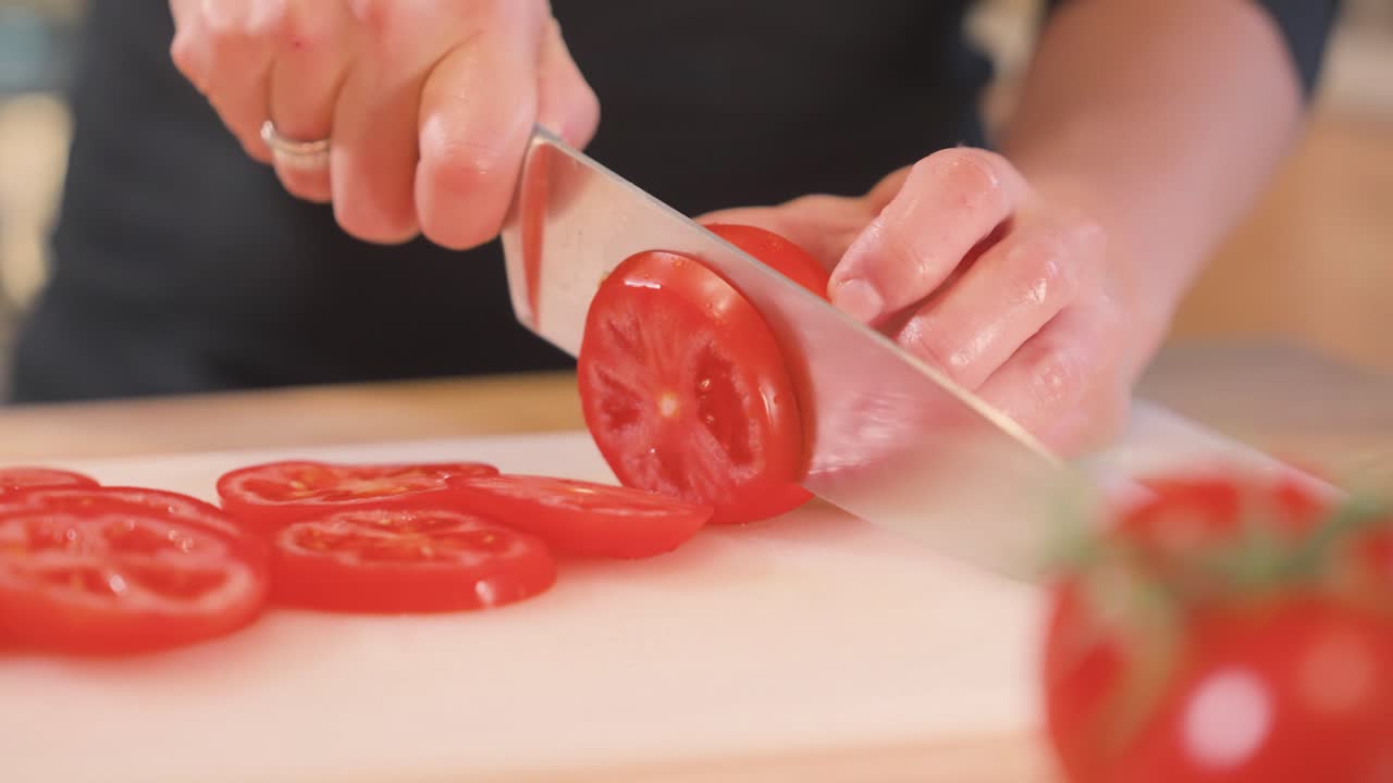 An at-home cook slices a tomato on a cutting board using a chef’s knife.