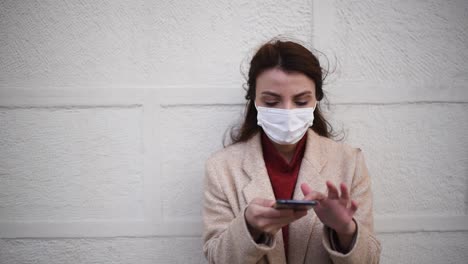 slow motion: beautiful girl wearing protective medical mask and fashionable clothes speaks with a smart phone in front of wall