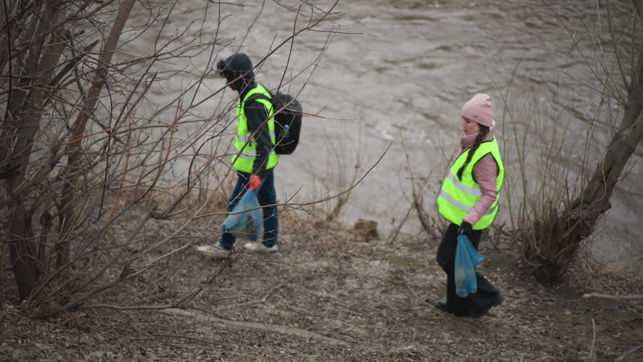Volunteer in reflective vest carrying blue plastic bag filled with collected litter walking along riverbank during environmental cleanup, partially obscured by large tree trunk in foreground