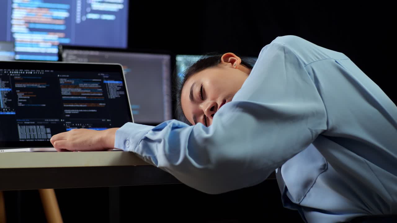Close Up Back View Of Asian Female Programmer Sleeping While Writing Code By A Laptop Using Multiple Monitors Showing Database On Terminal Window Desktops In The Office