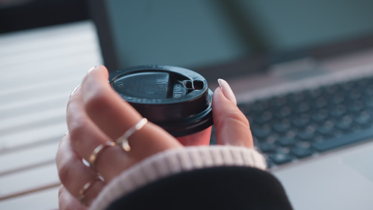 Close-up of woman hand gently turning coffee cup with manicured fingers, laptop blurred in background creating a sophisticated workplace vibe