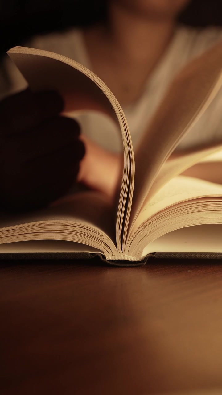 La mano di una donna gira le pagine di un libro aperto appoggiato su un tavolo di legno scuro, immerso nella calda luce dorata di una lampada, creando un'atmosfera di lettura intima e accogliente con un morbido bokeh sullo sfondo.