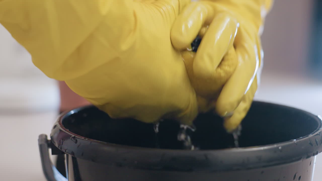 Close up of person wearing yellow gloves holding rag above black bucket on wooden floor with blurred robotic vacuum in background, household cleaning scene showing hygiene