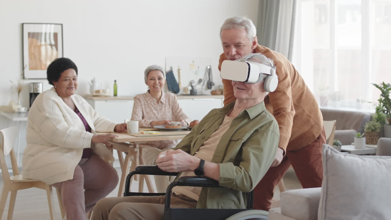 Medium long of happy handicapped man wearing VR goggles with earphones, his senior Caucasian friend moving wheelchair sideways, women looking at them smiling on background