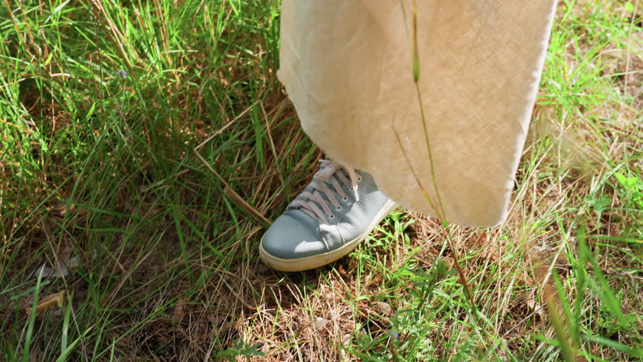 Aerial view of lady wearing light gown and blue jacket standing in grassy field under sunlight, surrounded by lush greenery, showing gentle movement, and natural simplicity of outdoor life