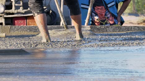 Construction-workers-pulling-mixed-cement-and-gravel,-spreading-it-over-a-floor-layout-of-a-building-under-construction-in-Southeast-Asia