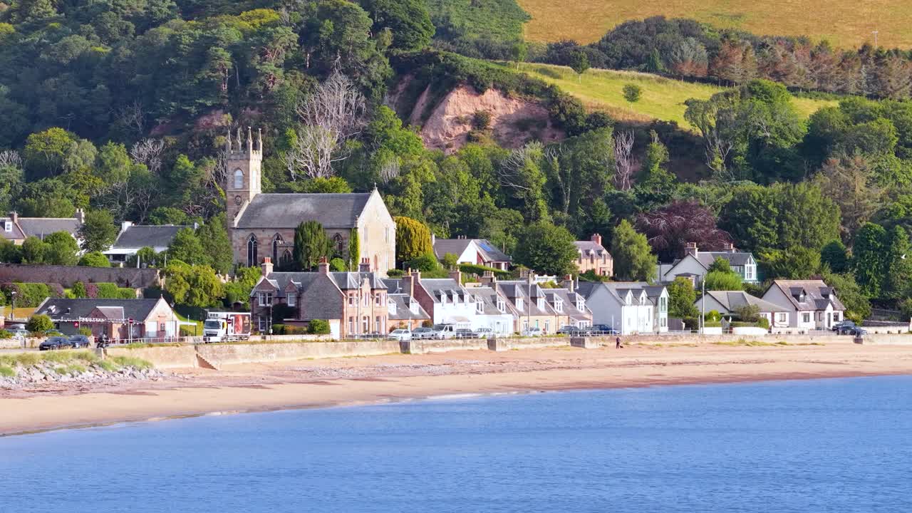 Daytime wide shot pans over Rosemarkie’s coastal village, historic church, greenery, and sandy shoreline