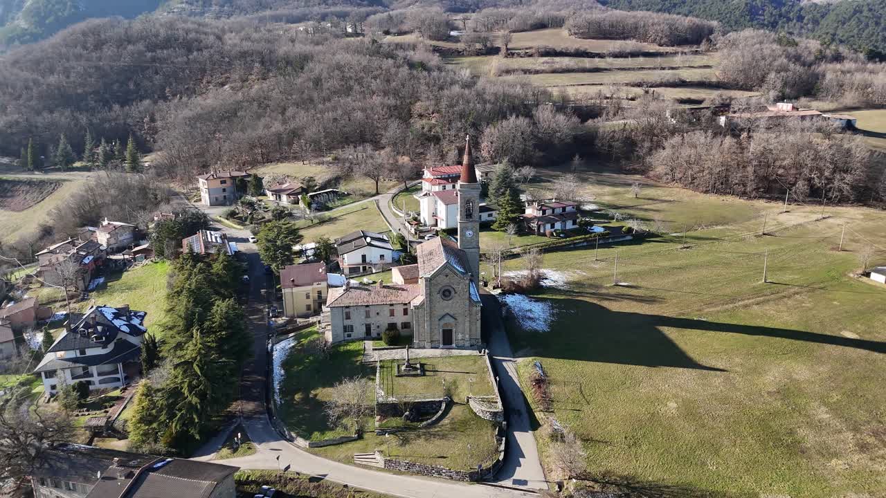 Zvonice Santi Salvatore e Gallo tyčící se nad klidnou I Rabbini, Morfasso PC, tradiční kamenný kostel a domy uhnízděné v zelených Apeninských údolích pod jasnou slunečnou oblohou, orbitální dron, Itálie