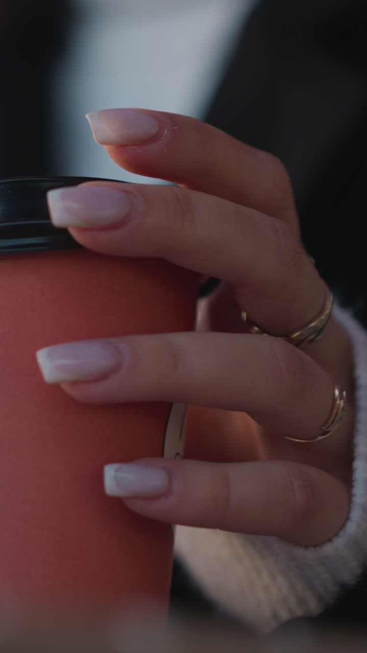 primer plano de la mano de una mujer con uñas cuidadosas tocando ligeramente la tapa de una taza de jugo roja mientras un fondo borroso revela un entorno profesional