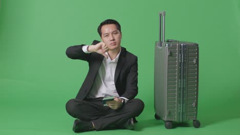full body of asian businessman traveler with luggage and passport looking at camera and showing thumbs down gesture in the green screen background studio