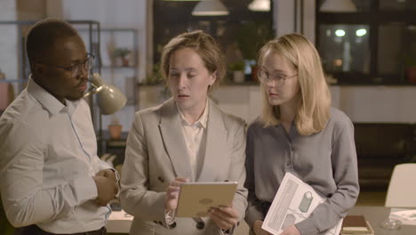 group of three  employees standing in the office, analyzing data on a tablet and discussing marketing strategy together 1