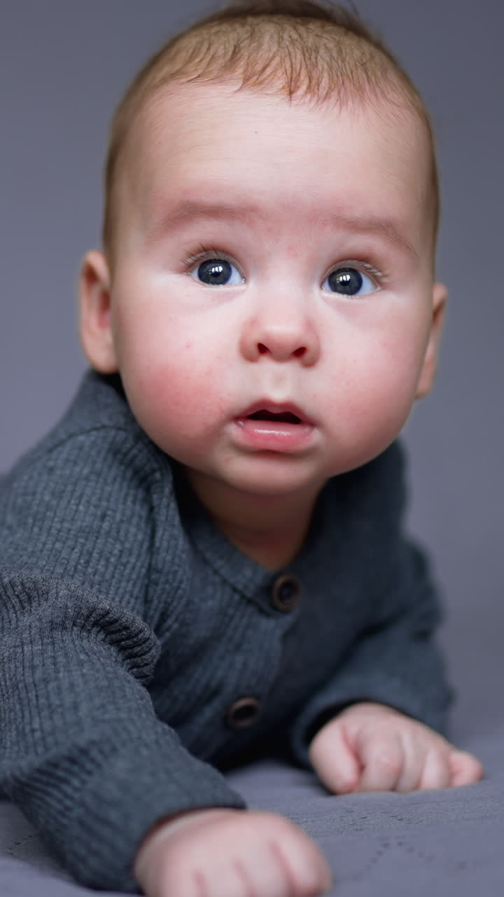 Healthy lovely kid in grey clothes lies on his belly. Adorable child looking sideways with interest. Grey blurred backdrop. Vertical video