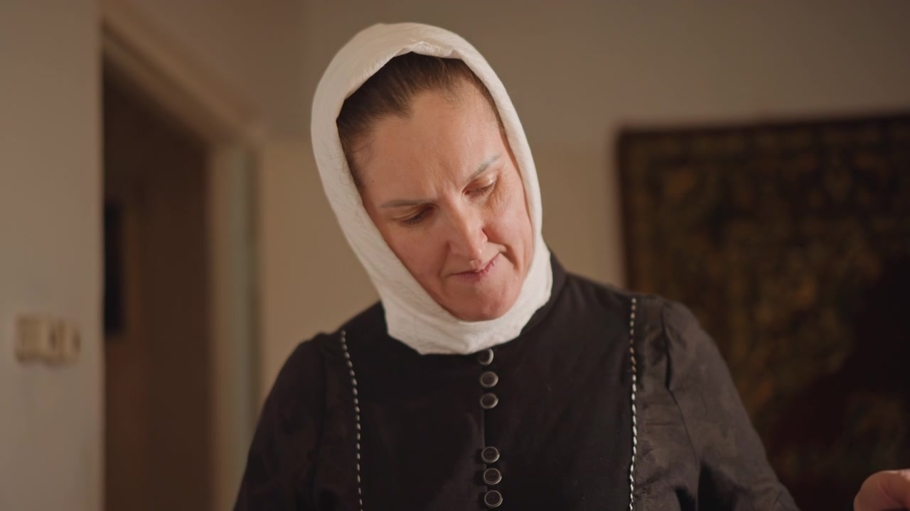 Vrouw met witte muts kijkt naar beneden in een warm verlicht vintage interieur, gekleed in een eenvoudige donkere jurk met knoopdetails, terwijl ze een rustige huishoudelijke taak uitvoert, zoals naaien of stof vouwen. Close-up beelden vangen de zachtheid van het moment.