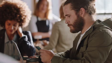 joven caucásico usando una computadora portátil en un café navegando en línea revisando mensajes en su teléfono inteligente trabajando en correos electrónicos en un restaurante ocupado