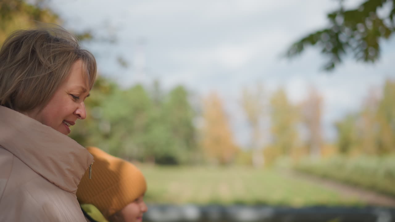 Woman walks side by side with son along leaves strewn forest path, both looking down at dog leash in hand while enjoying crisp autumn day under canopy of golden trees